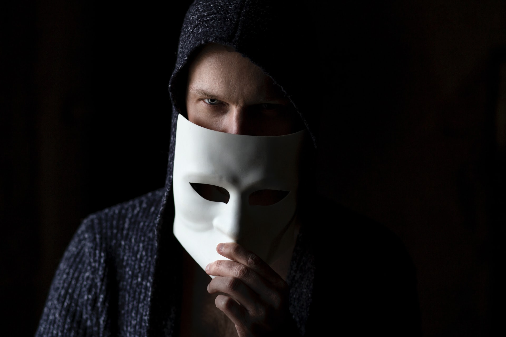a man in a black hood against a black background with a white mask half covering his face looking sinister